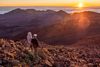 Two people stand on rocky terrain, watching the sun rise over layered mountain peaks.