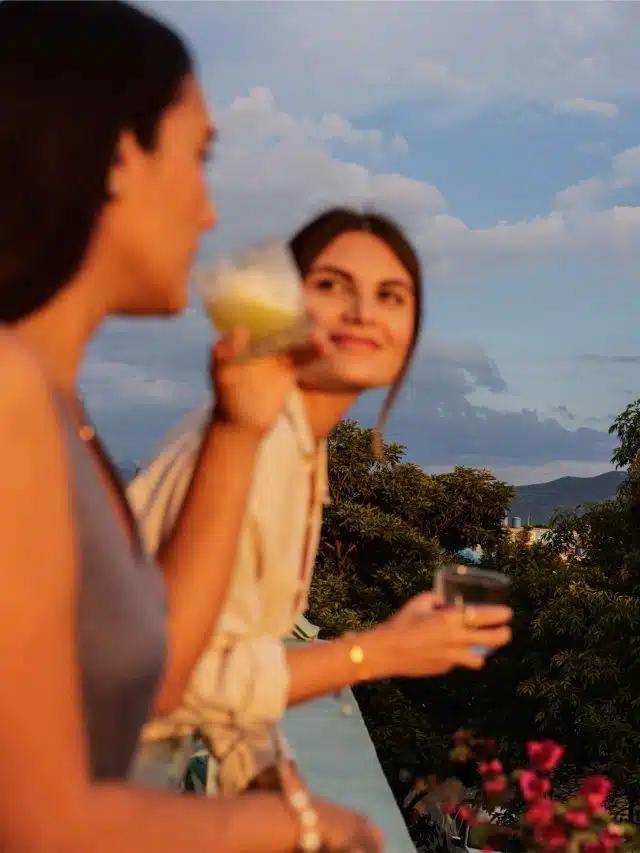 Two women stand on a balcony holding drinks with trees and flowers below in the distance