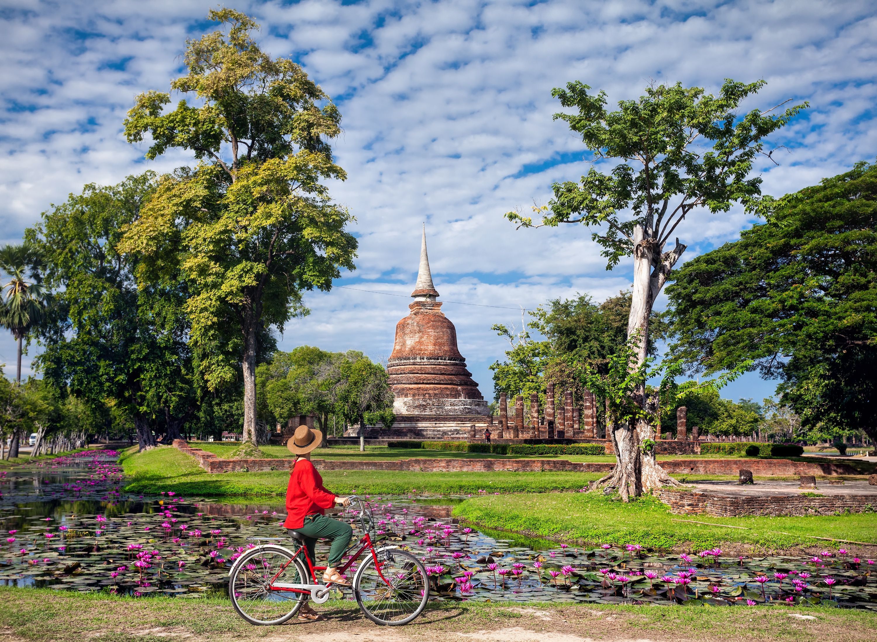 A woman wearing a straw hat and wearing a red bicycle pauses along a pathway to look at Wat Sa Si temple in the lush, nature-filled Sukhothai Historical Park in Thailand