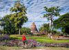 A woman wearing a straw hat and wearing a red bicycle pauses along a pathway to look at Wat Sa Si temple in the lush, nature-filled Sukhothai Historical Park in Thailand