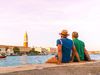 Two men sit along along a river overlooking city buildings.