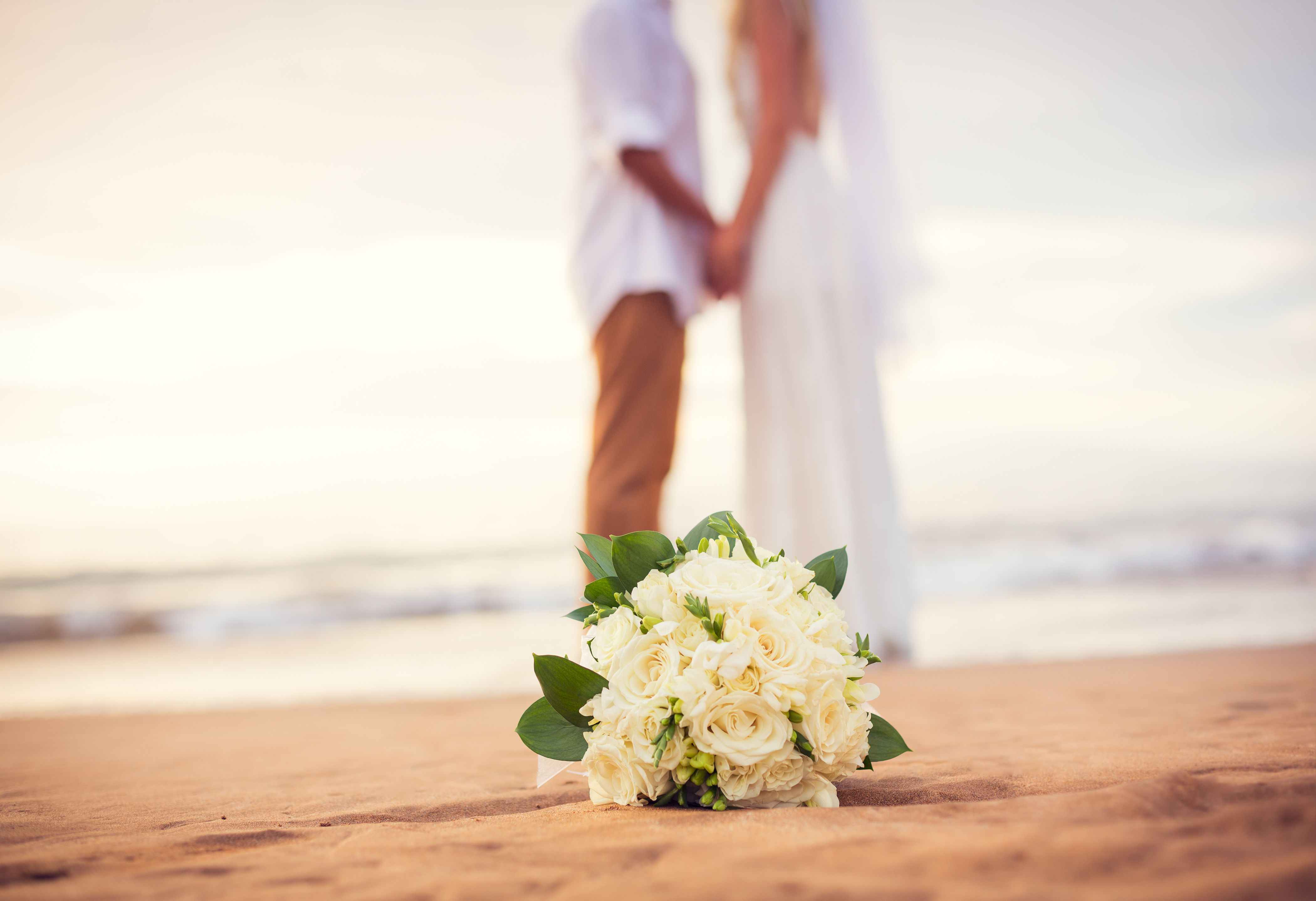 A beach wedding ceremony on a beach, featuring a couple in the background and a bridal bouquet resting in the sand.