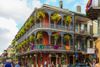 A colorful, balconied corner building in New Orleans' French Quarter, with flags, and ferns hanging from elaborate ironwork