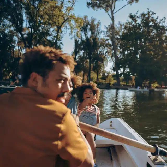 A man and a young girl are in a rowboat on the lake at Villa Borghese in Rome, Italy, on a sunny day with trees framing the lake in the background. She is excitedly pointing at something behind the camera and he has turned his head to look