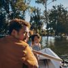 A man and a young girl are in a rowboat on the lake at Villa Borghese in Rome, Italy, on a sunny day with trees framing the lake in the background. She is excitedly pointing at something behind the camera and he has turned his head to look