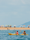 Two people kayak in the ocean in front of a golden sand beach