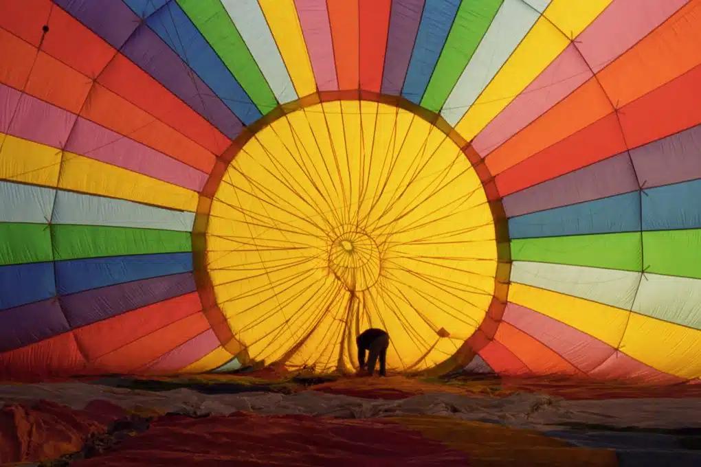 A person stands inside the envelope of a rainbow-colored hot-air balloon as it inflates