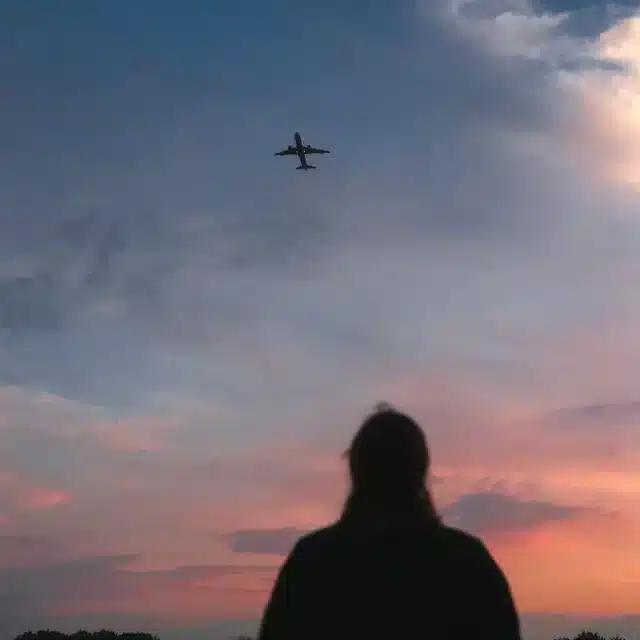 A silhouette of a figure standing before a pink cloudy sky and looking up at a plane passing overhead