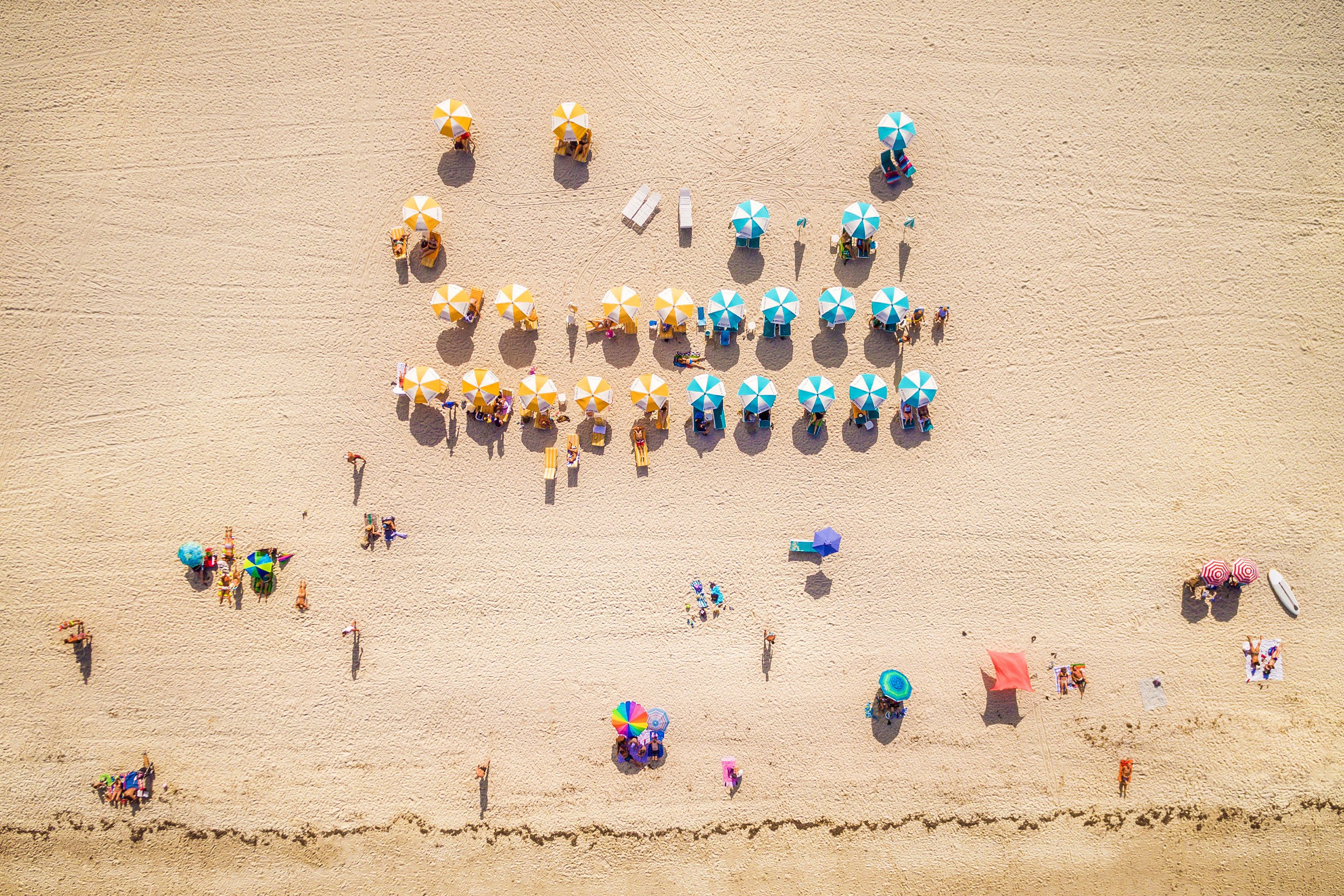 Overhead view of yellow and blue beach umbrellas and people laying in the sun on a beach in Miami.