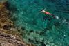 A person wearing orange swim trunks snorkels in the clear water at the edge of a rocky bay