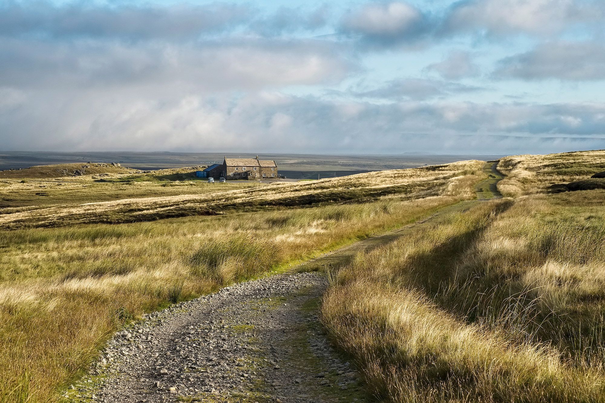 A pub stands alone in the middle of a landscape with a rocky trail in the foreground in Reeth, England.