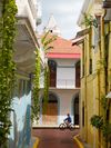 View down a narrow street in Casco Antiguo, Panama City, with pastel color buildings and a cyclist passing