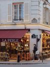 La Perle restaurant on a street corner in Paris, France, with sidewalk seating and a shopper walking past