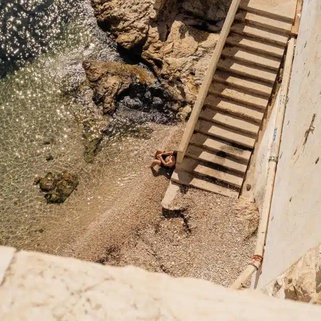 A person sits on a pebbly beach with their feet in the sea, at the base of stone steps, in Marseille, France