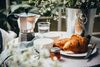 Softly blurred flowers in the foreground frame a plated croissant on a table with cups and craft.