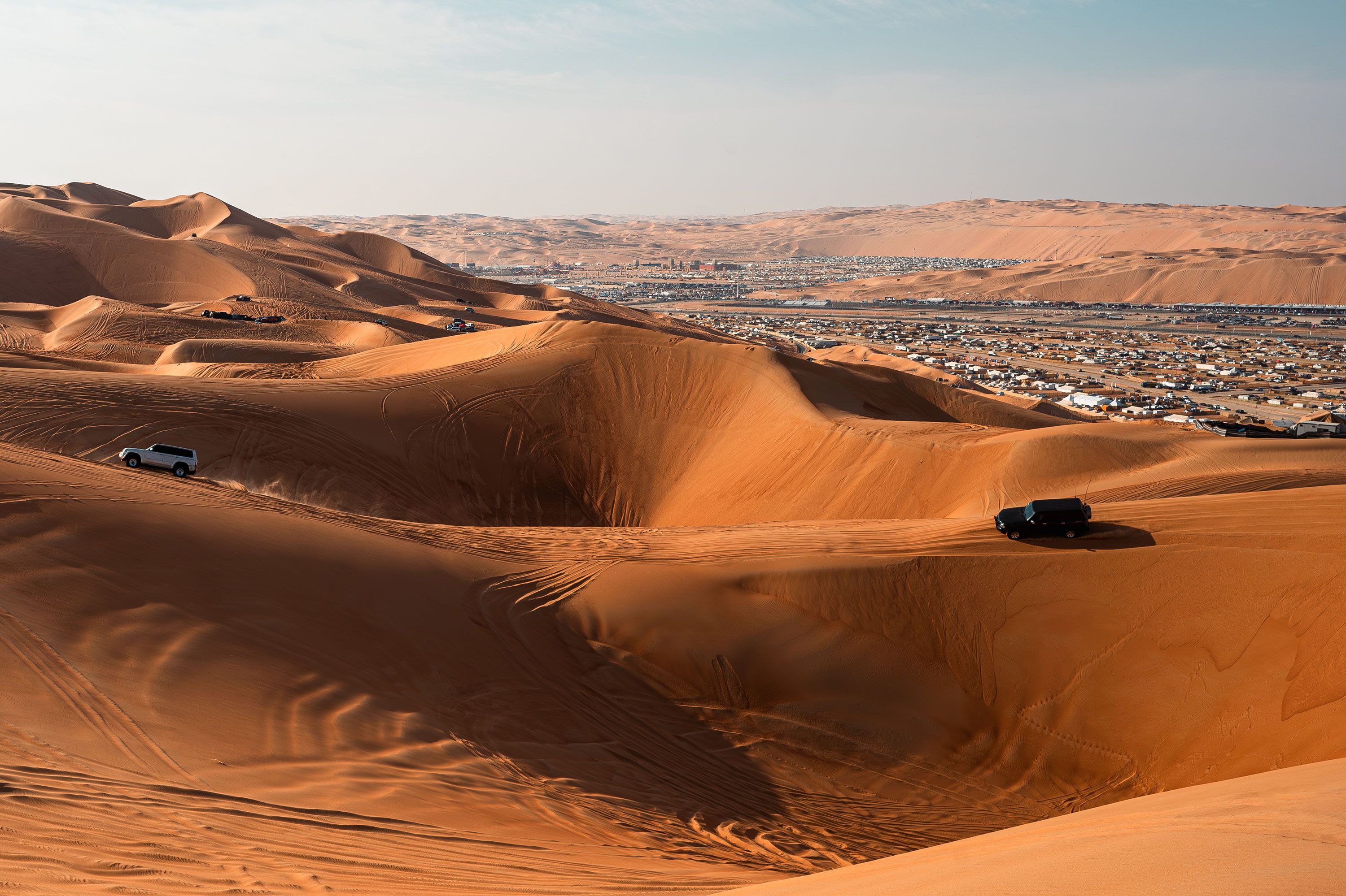 A wide desert landscape in Abu Dhabi with golden sand dunes and faint tire-tracks winding toward a small encampment visible on the horizon.