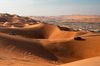 A wide desert landscape in Abu Dhabi with golden sand dunes and faint tire-tracks winding toward a small encampment visible on the horizon.