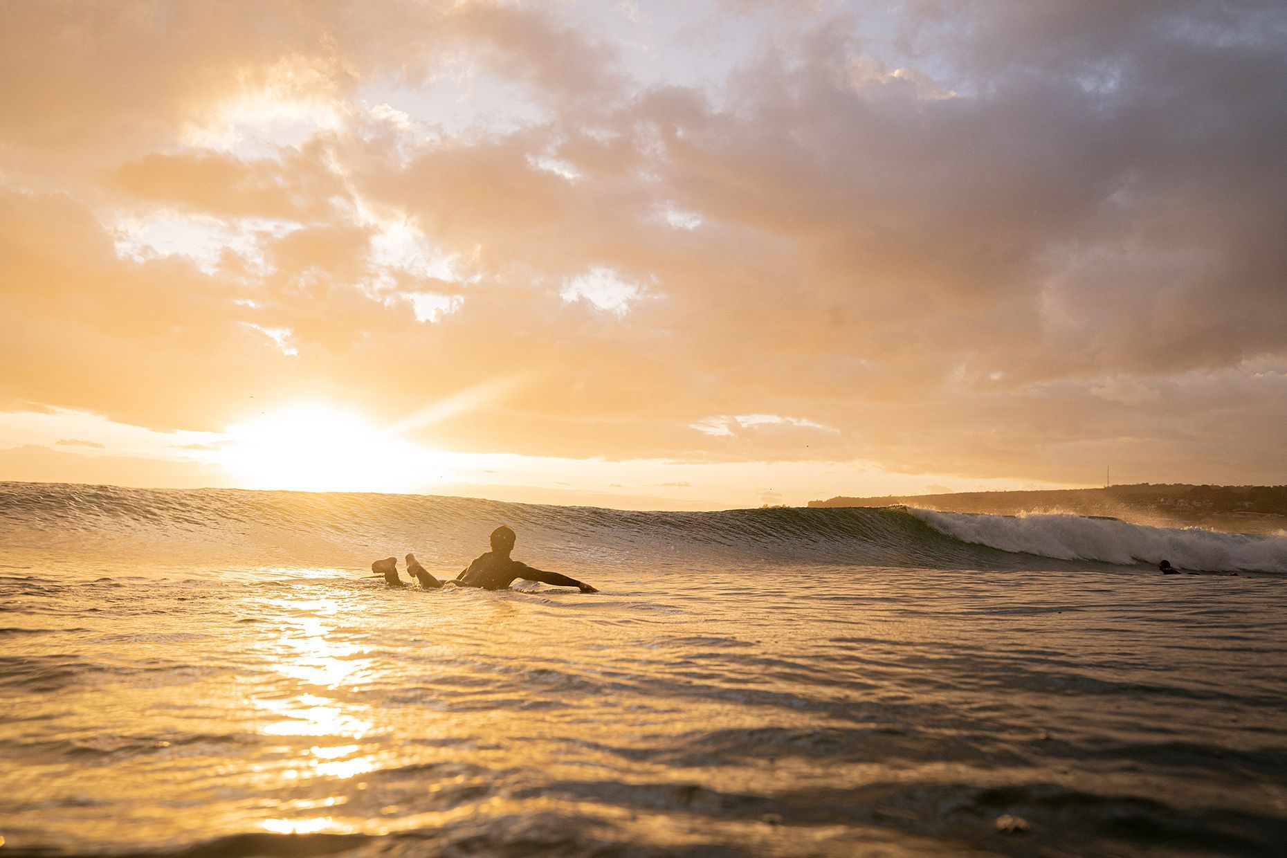 A surfer paddles out as a wave crests, the sun rising over the water