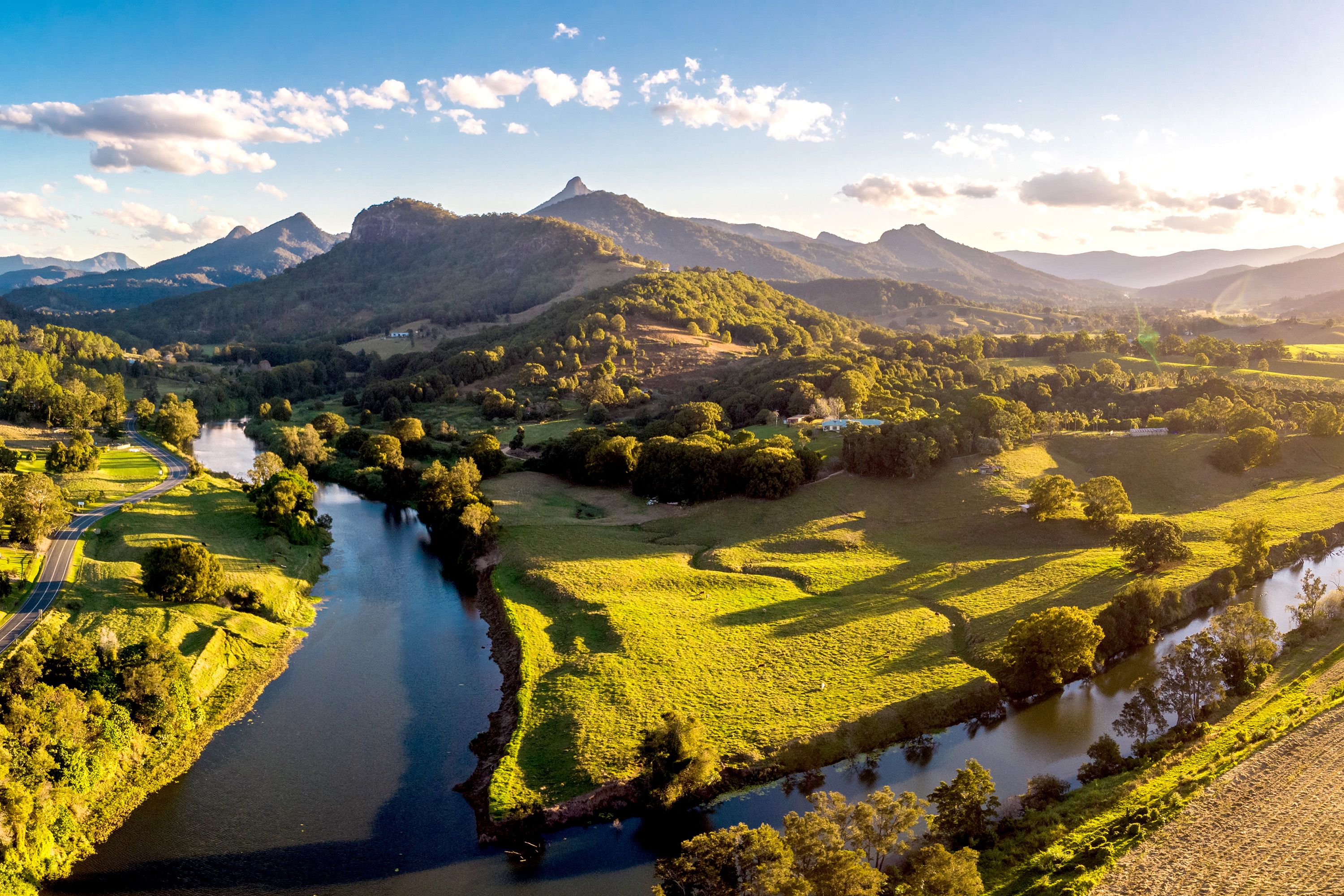 River forks between grassy fields and trees with mountains in the distance.