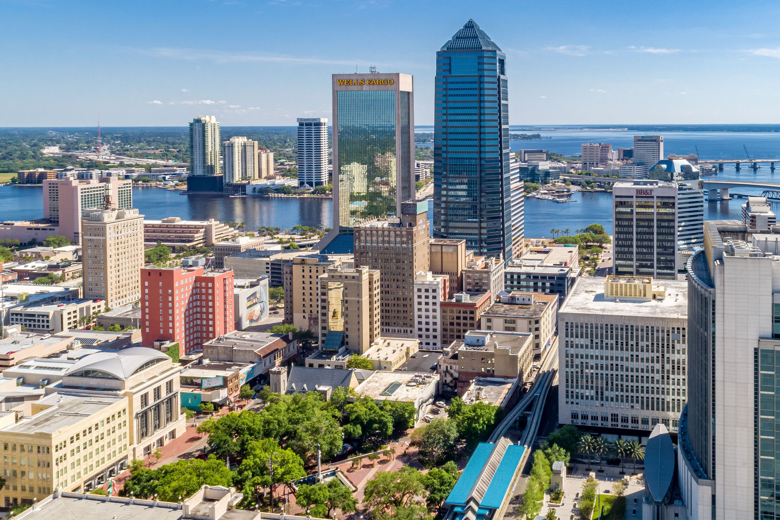 A wide-angle image of Jacksonville, Florida showing city skyscrapers along the St. Johns River, a bridge span, and the coastline in the distance.