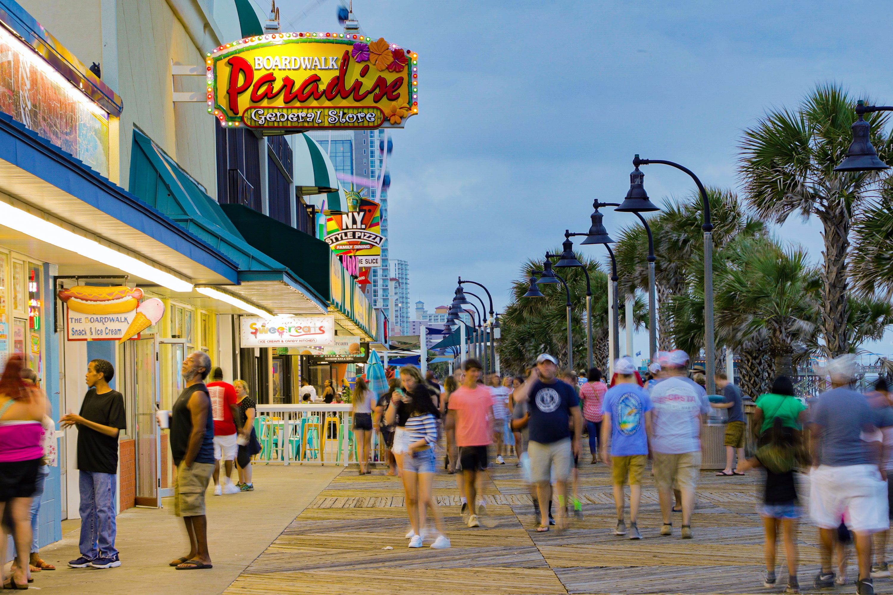 People walking along the Myrtle Beach Boardwalk with illuminated storefronts at dusk.