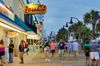People walking along the Myrtle Beach Boardwalk with illuminated storefronts at dusk.