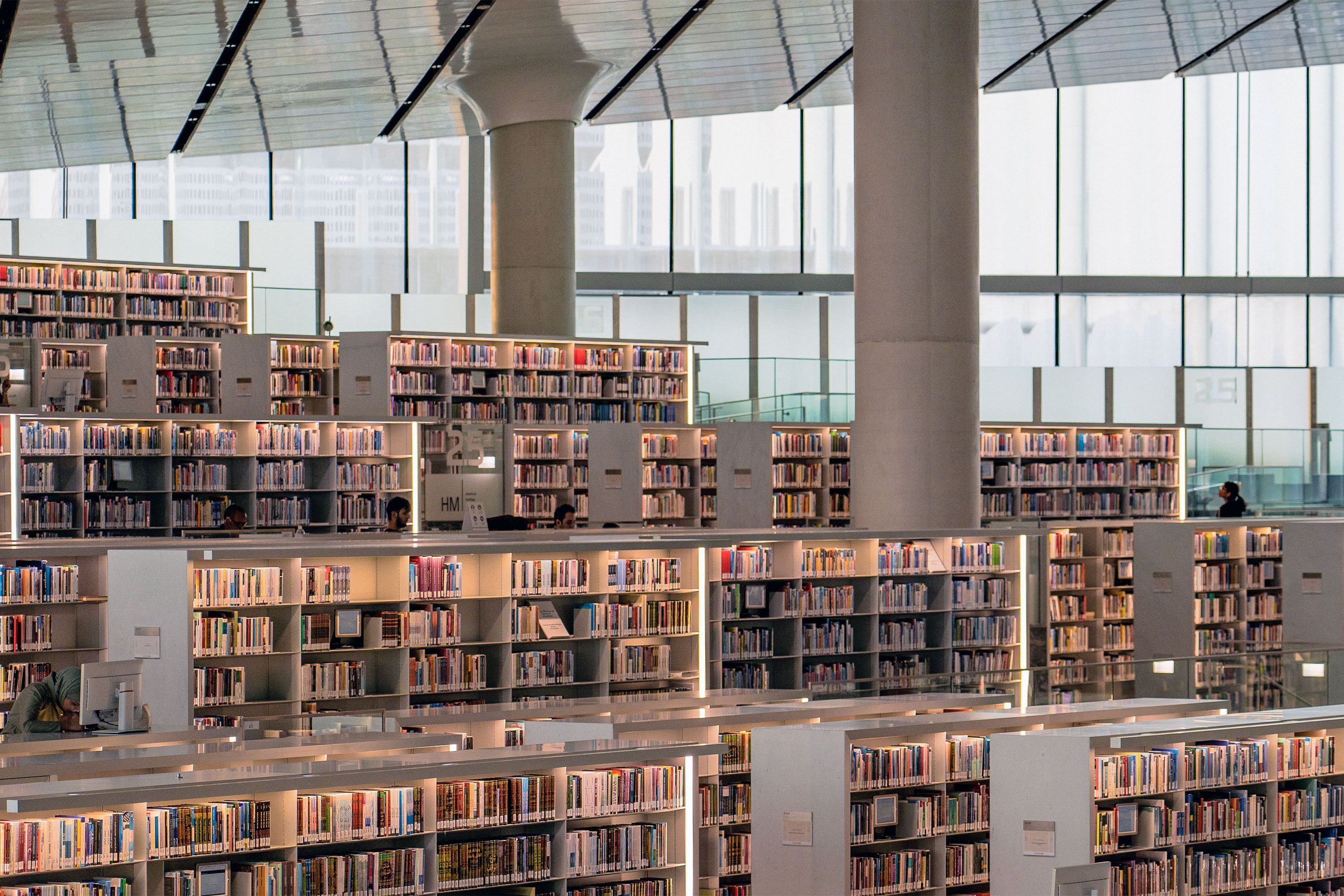 An interior view of the Qatar National Library showing rows of white bookshelves, wide open walkways, and natural light streaming through large glass walls in a contemporary architectural setting.