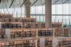 An interior view of the Qatar National Library showing rows of white bookshelves, wide open walkways, and natural light streaming through large glass walls in a contemporary architectural setting.