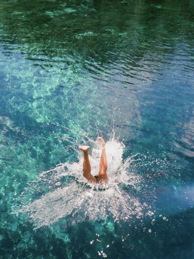 A person's legs are seen from above the surface as they plunge into calm turquoise waters