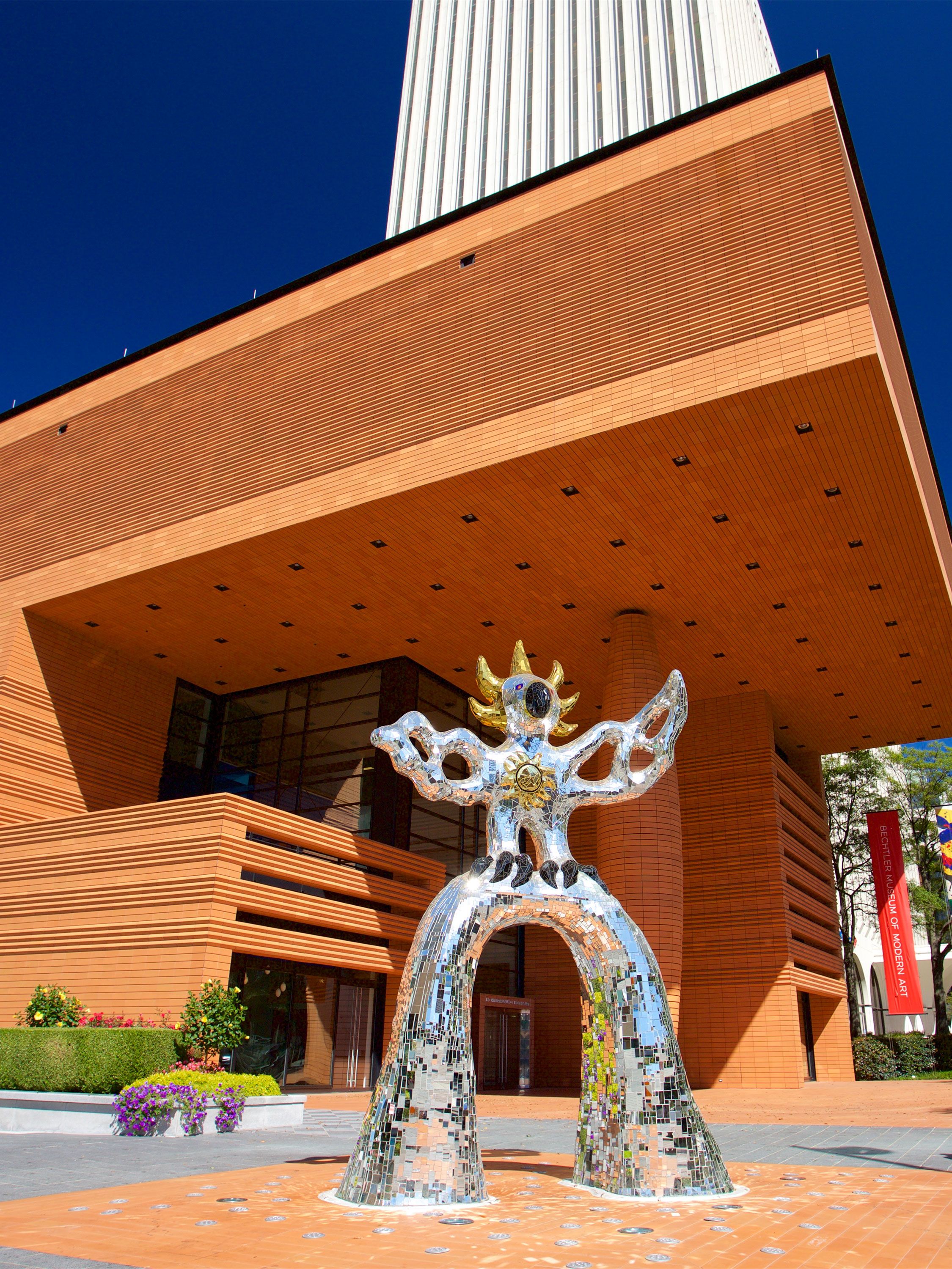 A mirrored sculpture with outstretched arms stands outside a modern museum entrance.