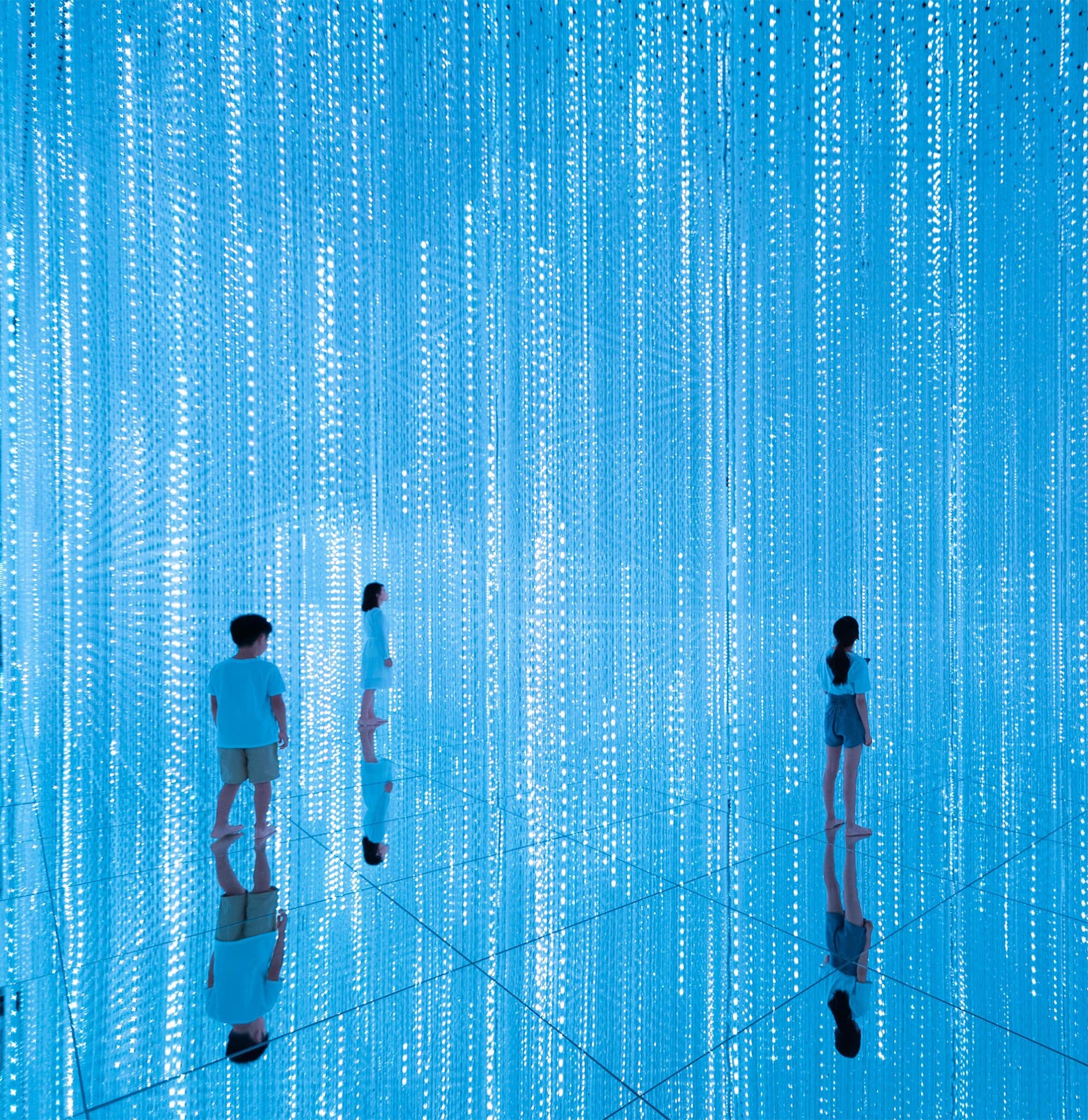 Visitors stand in a bright blue room, with vertical strips of sparkling lights, at the teamLab's Infinite Crystal World, Tokyo