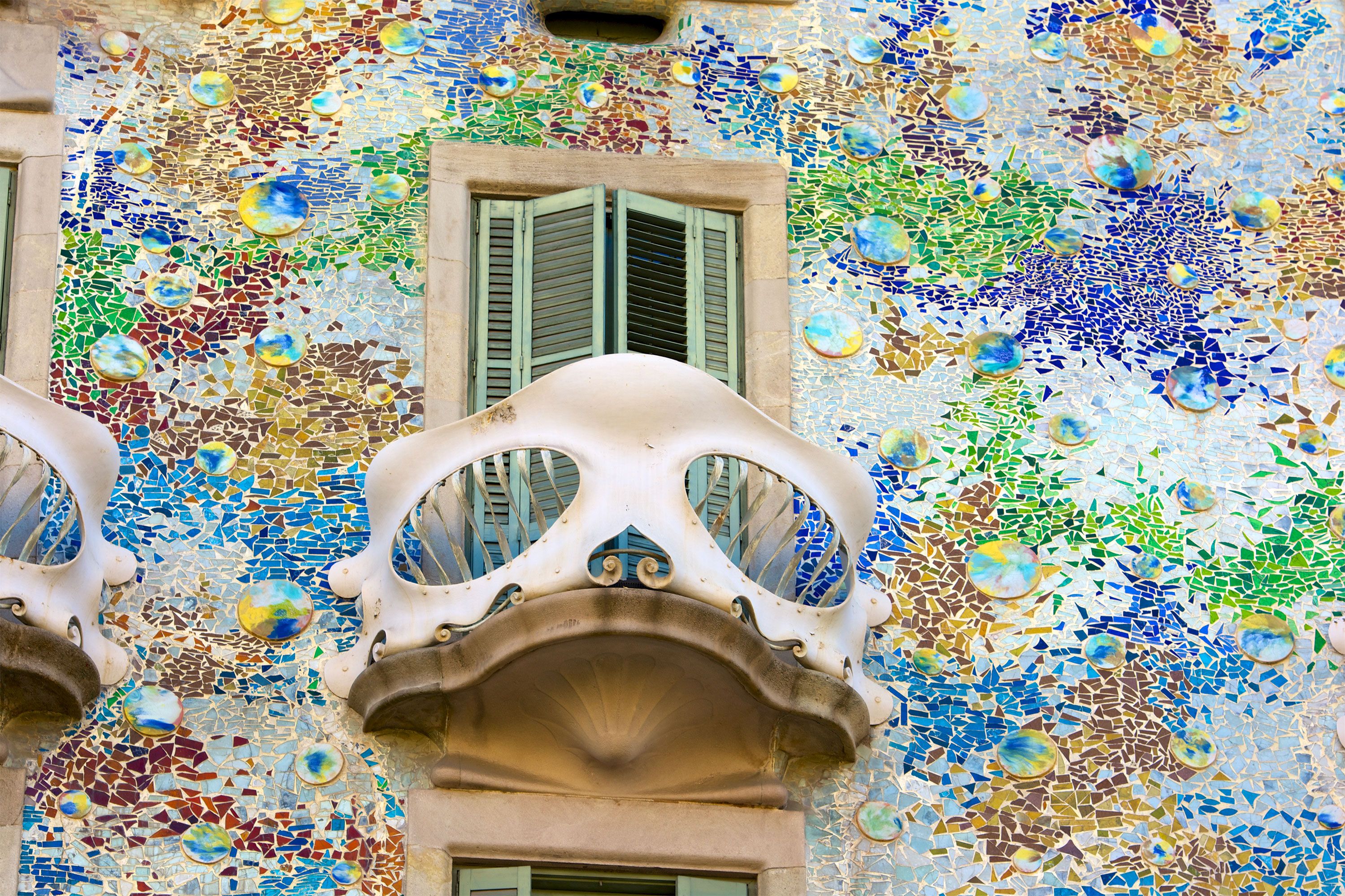 A close-up of a Gaudí-style building façade with a bone-shaped white balcony, green shutters, and colorful mosaic tiles in blues, greens, and golds.