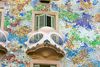A close-up of a Gaudí-style building façade with a bone-shaped white balcony, green shutters, and colorful mosaic tiles in blues, greens, and golds.