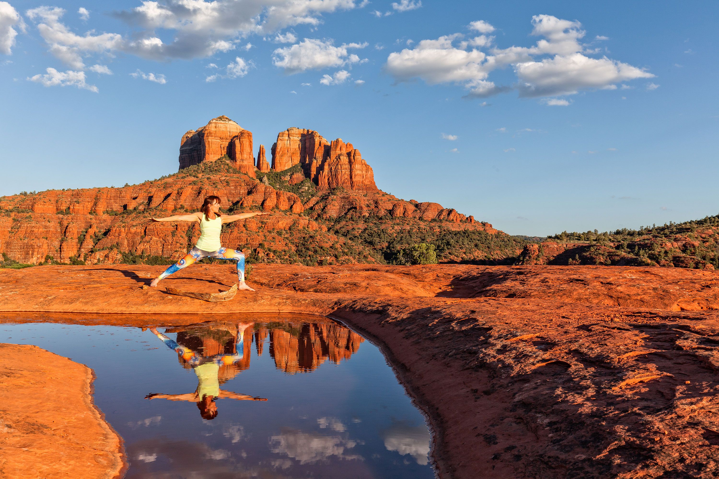 Woman does yoga in desert setting with red rock formations and reflexion in natural pool below blue cloudy sky.