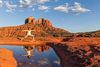 Woman does yoga in desert setting with red rock formations and reflexion in natural pool below blue cloudy sky.