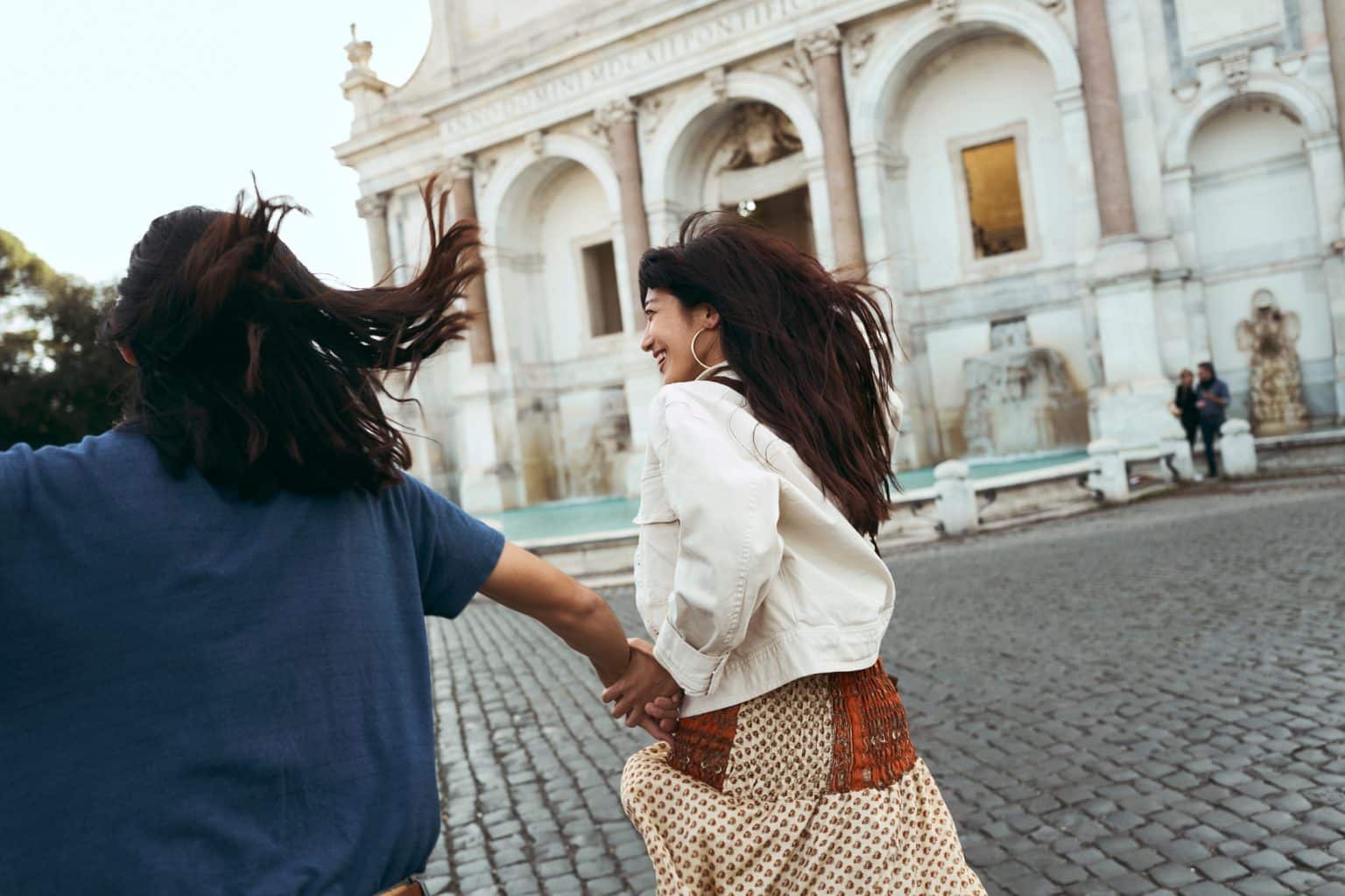 Two women run, laughing, through a square with a fountain in Rome