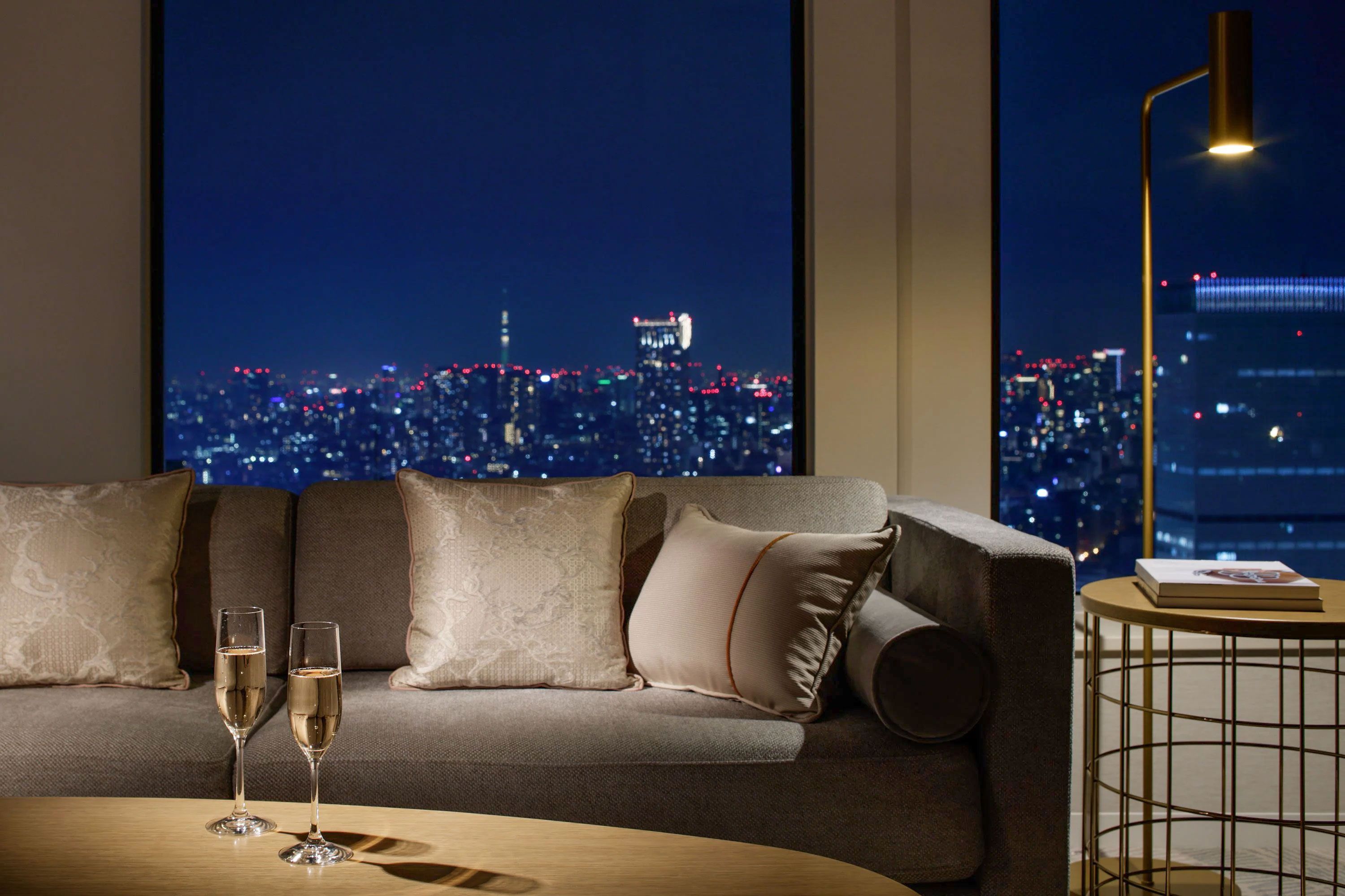 Two Champagne glasses sit on a coffee table in front of a couch with views of Tokyo at night in the background.