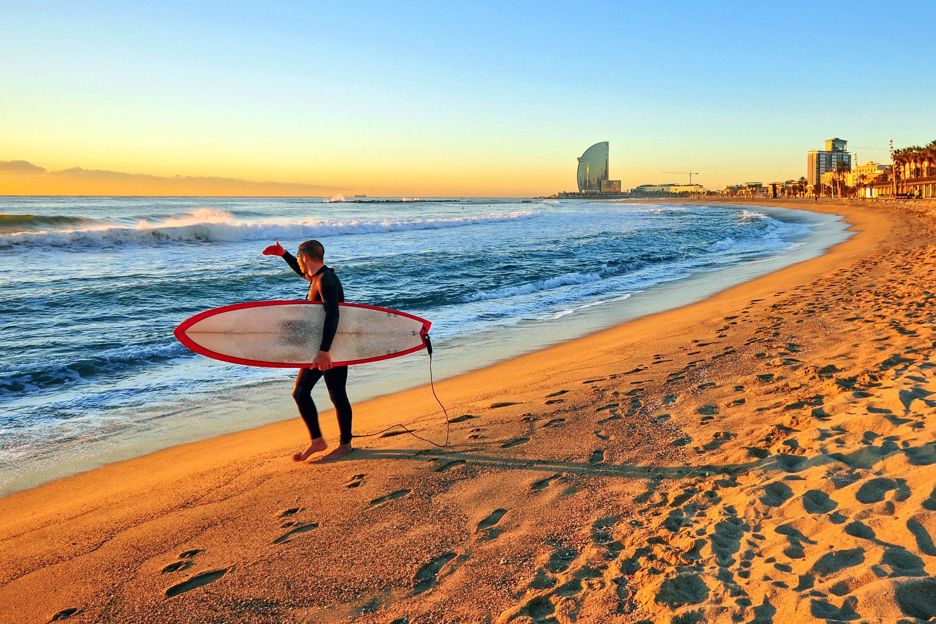 A surfer on a sandy beach shields his eyes from the sun at sunset as he looks out over the waves with a tall building in background.