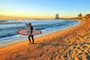 A surfer on a sandy beach shields his eyes from the sun at sunset as he looks out over the waves with a tall building in background.