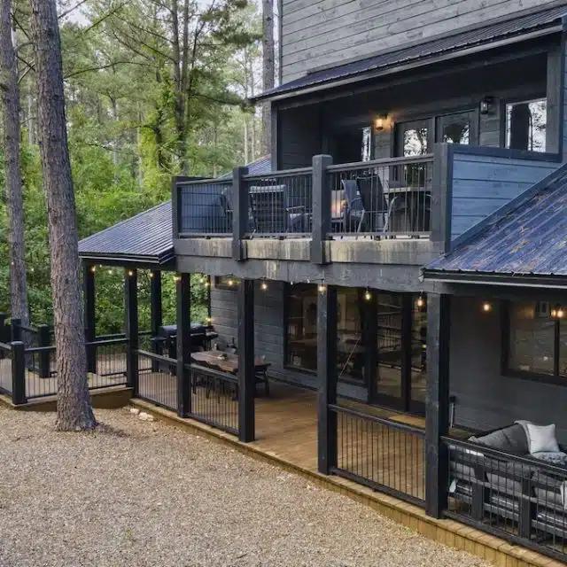 A black painted, wood paneled home in the woods, with a veranda on the upper floor