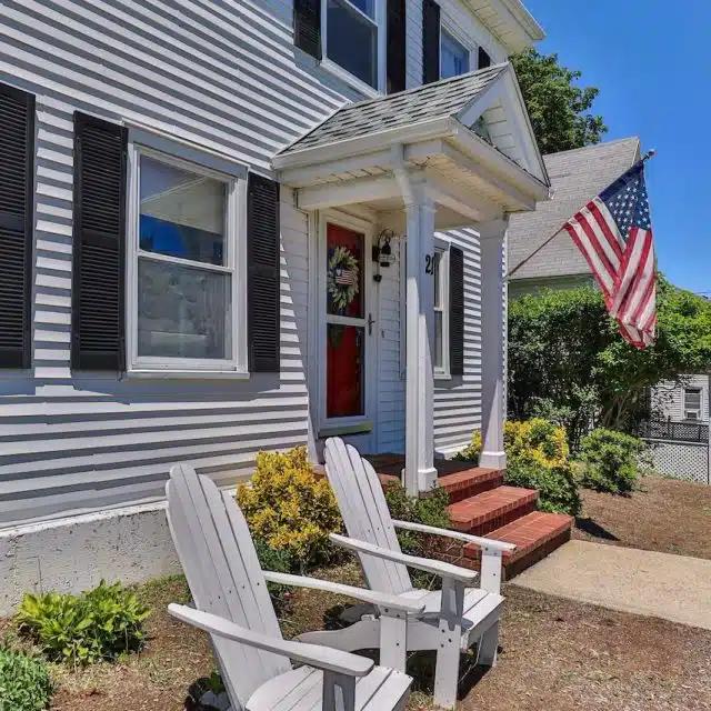 A white clapboard home with Stars and Stripes flag and Adirondacks in front