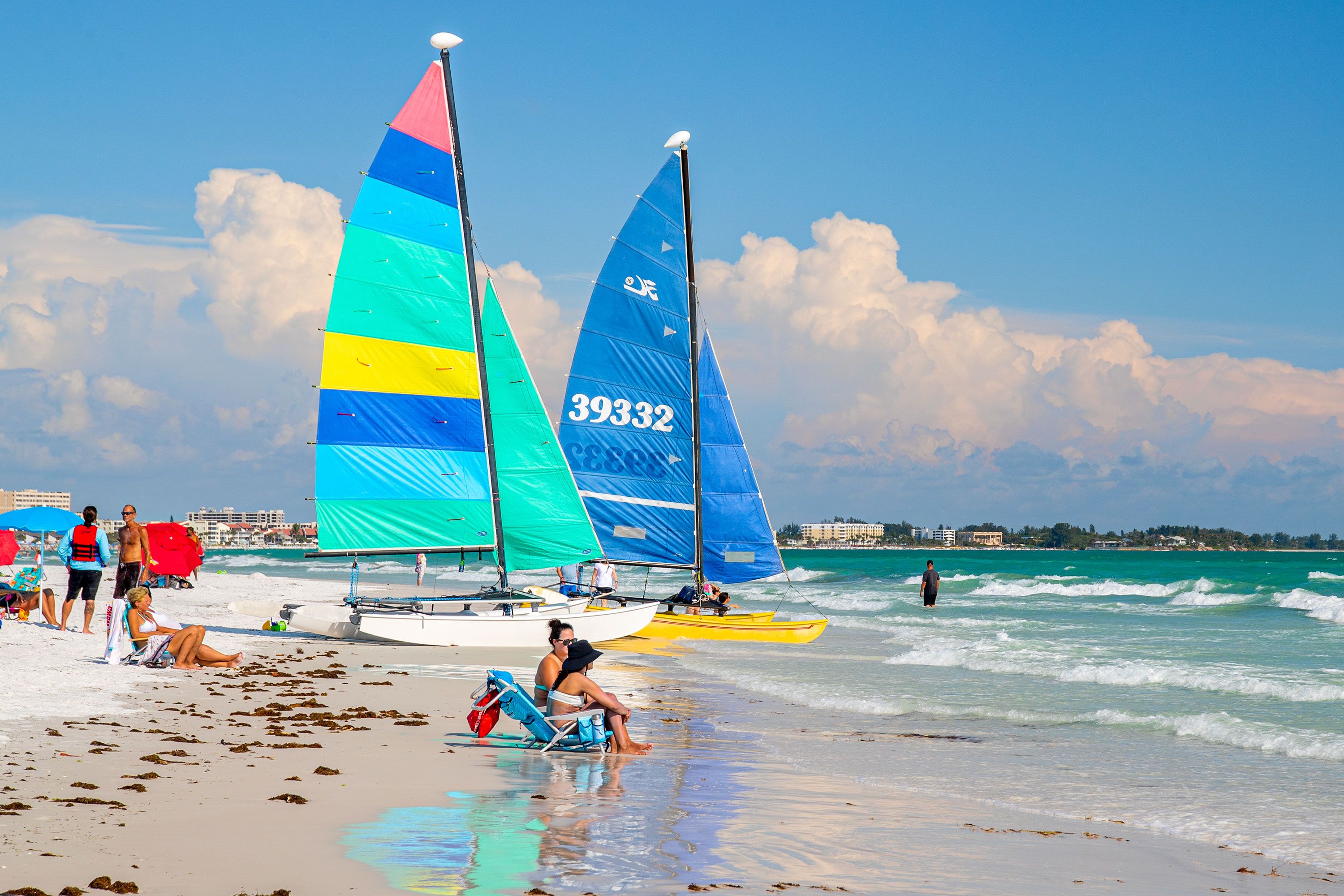 Brightly colored sailboats on a sandy beach with people sitting and wading in shallow ocean water.