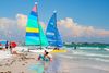 Brightly colored sailboats on a sandy beach with people sitting and wading in shallow ocean water.