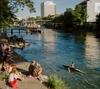 People sit on the steps at the edge of the river Limmat as someone kayaks past them on a sunny day in Zürich, Switzerland