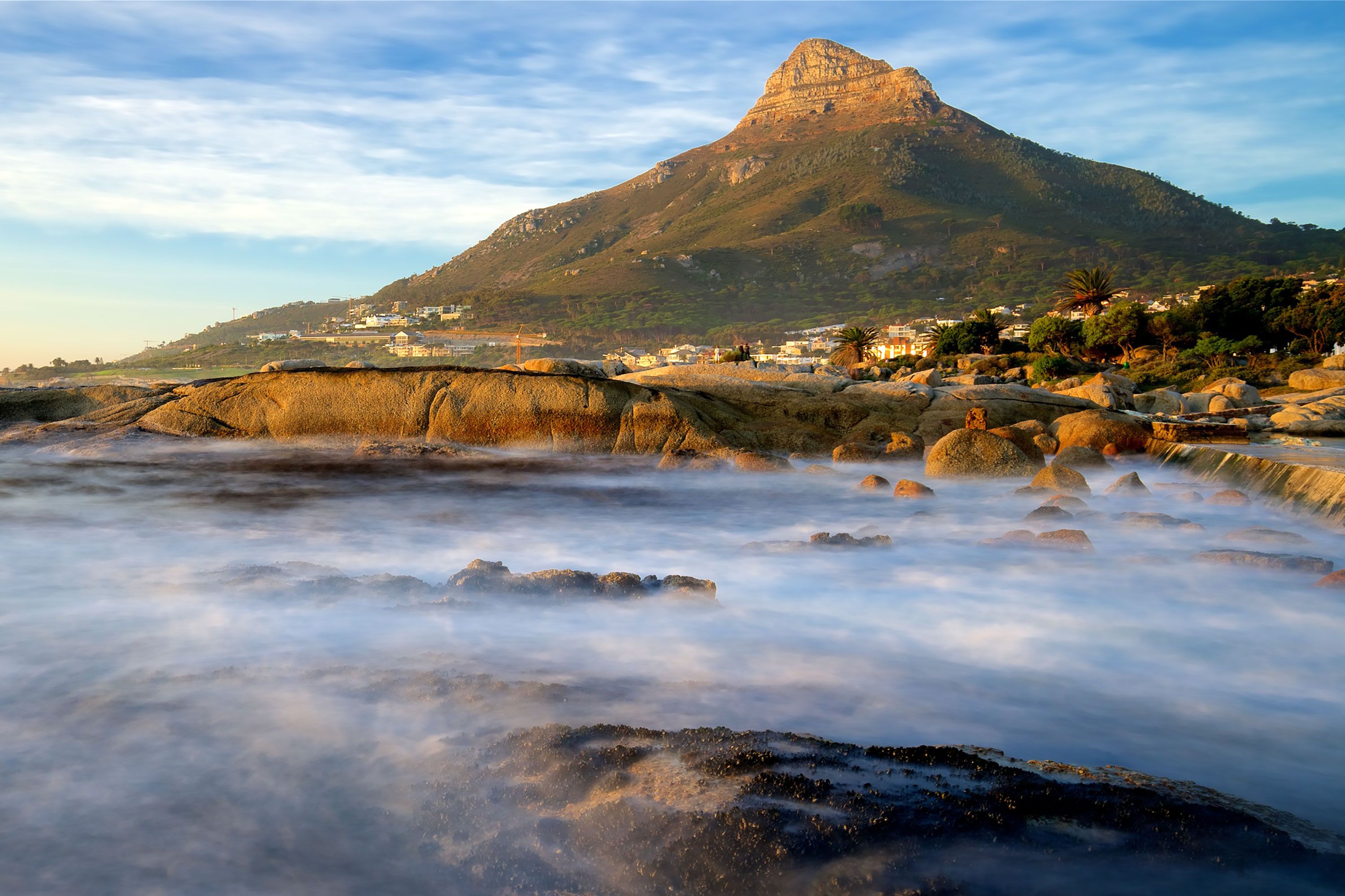 View of Lion’s Head mountain above Camps Bay with smooth, misty waves washing over rocks in the foreground at sunrise.