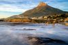 View of Lion’s Head mountain above Camps Bay with smooth, misty waves washing over rocks in the foreground at sunrise.