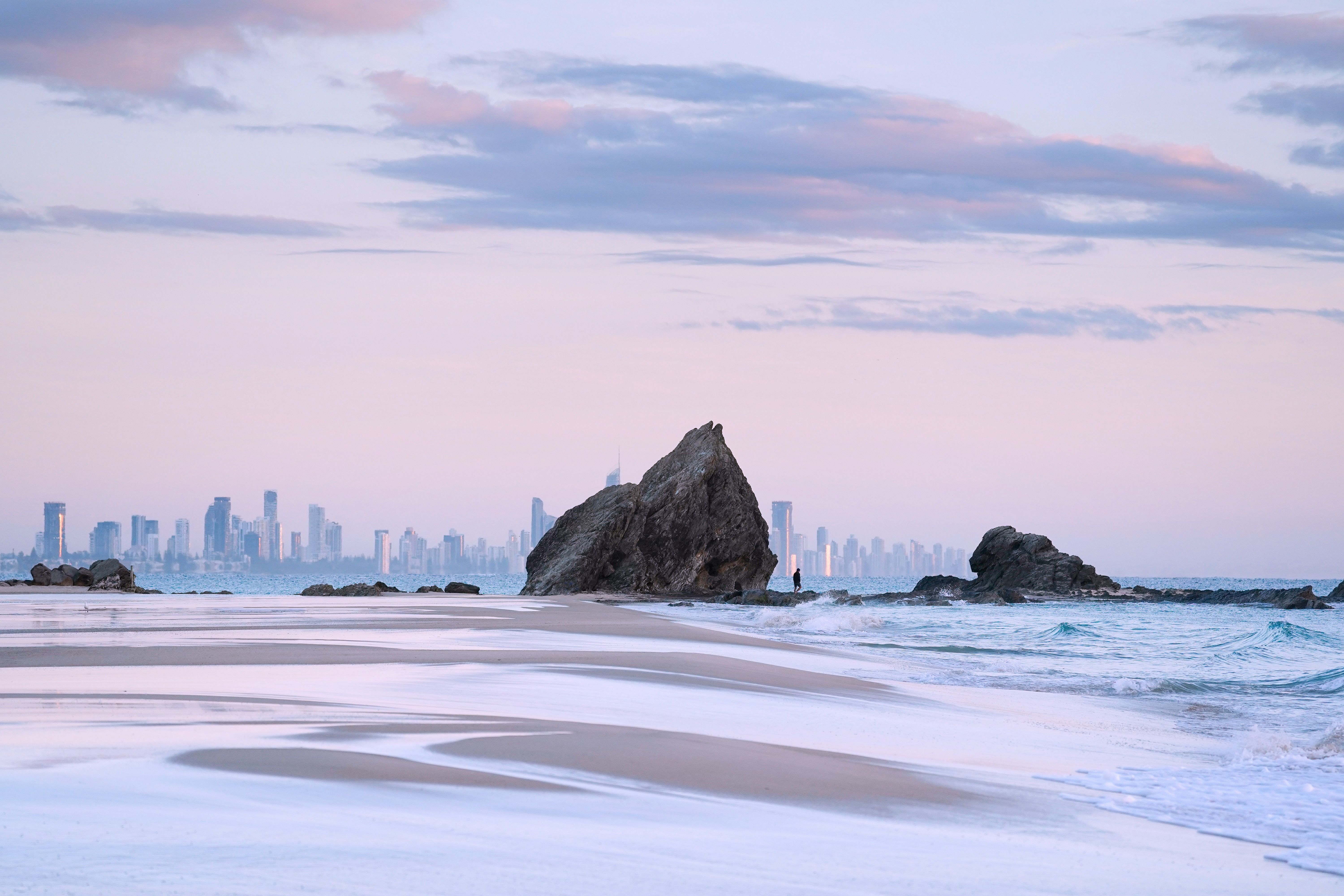 Large, craggy rocks rise out of the sand on Currumbin Alley beach with the Gold Coast skyline in the background