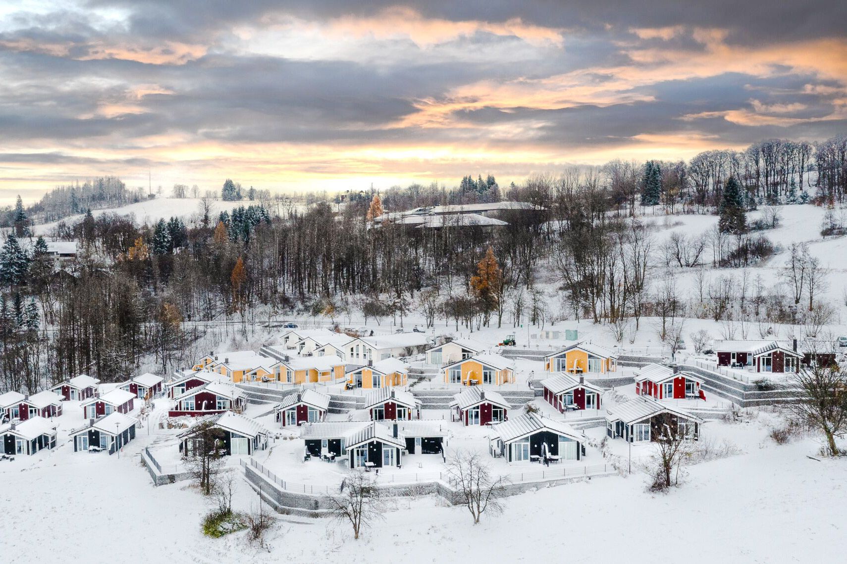 Eine Häusersiedlung in einer verschneiten Winterlandschaft, im Hintergrund ist ein Sonnenuntergang hinter den Wolken sichtbar