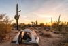 A man is sititng in a small domed tent in the desert with cacti at sunset.
