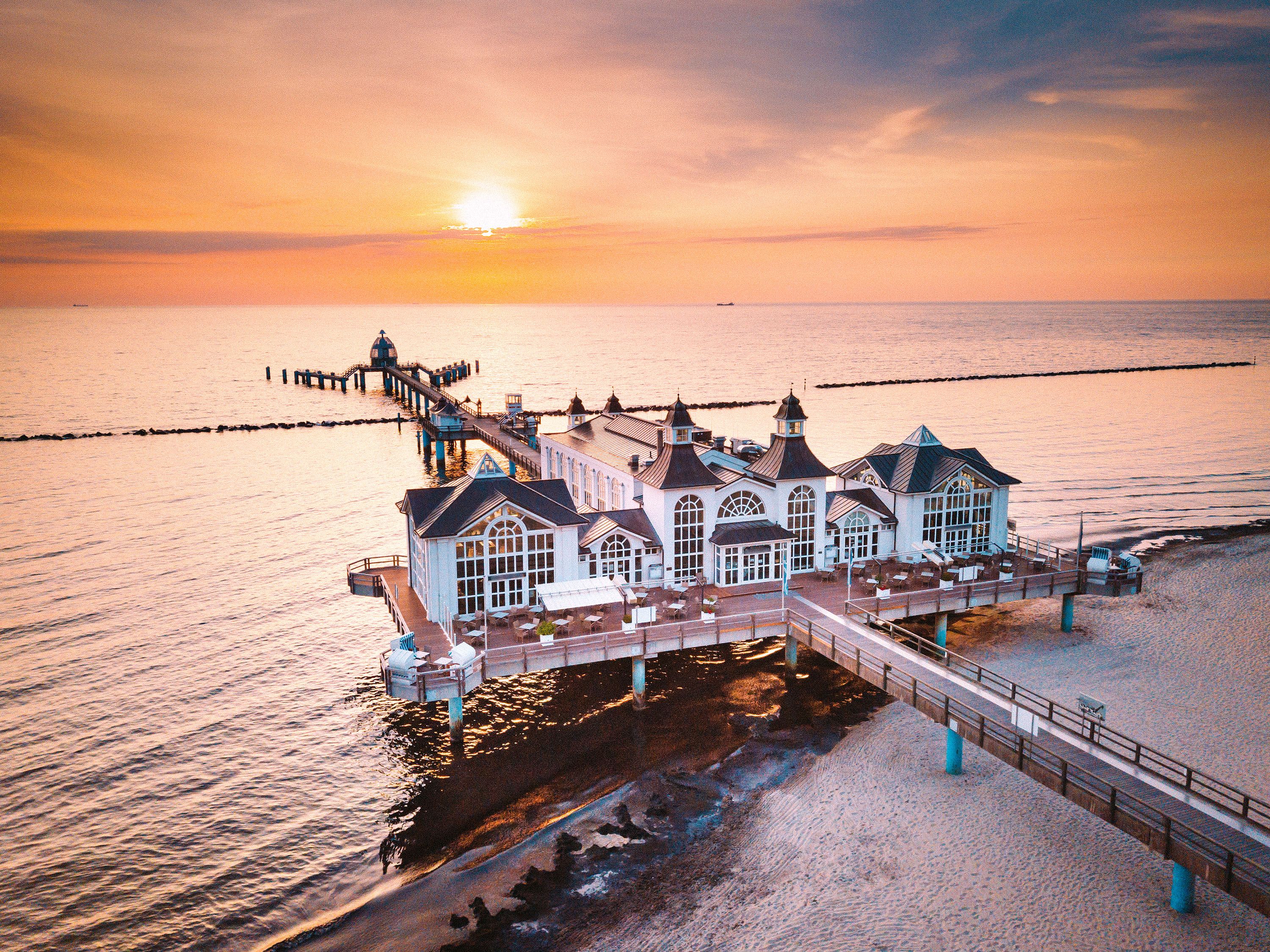 Aerial view of a large white building with many paned windows on a pier into calm waters.